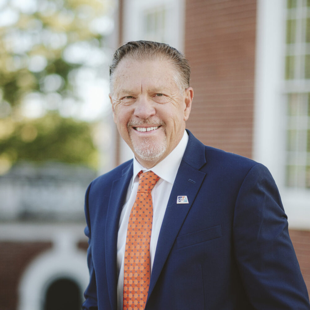 Jody Kielbasa in a navy suit with an orange patterned tie, smiling outdoors.