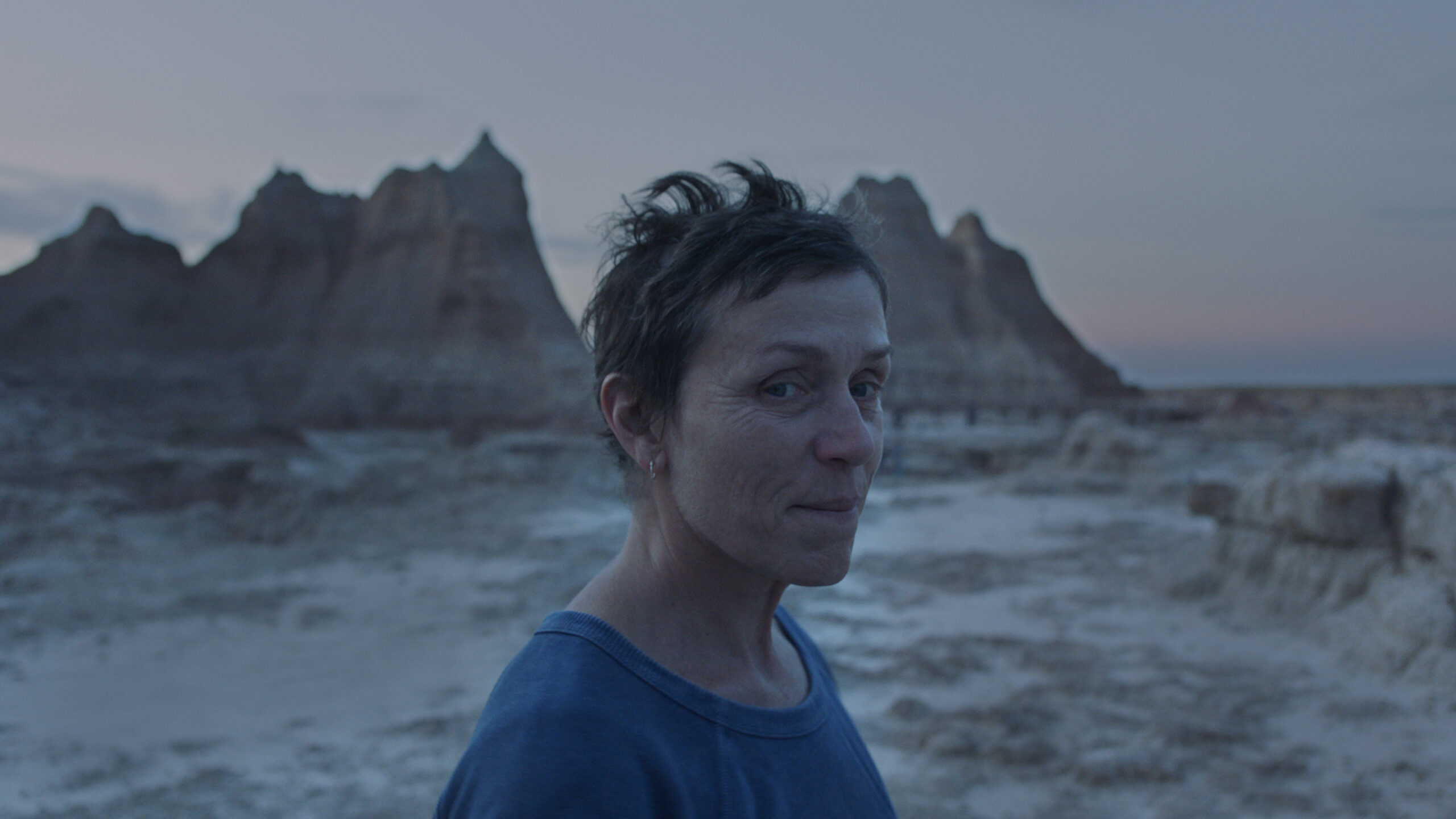 Frances McDormand in "Nomadland" against a backdrop of rugged rock formations at dusk.