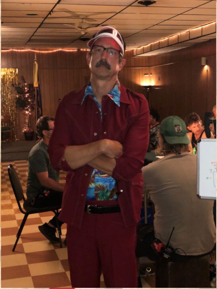 Brad DeMarea in a red suit, visor, and mustache on set.