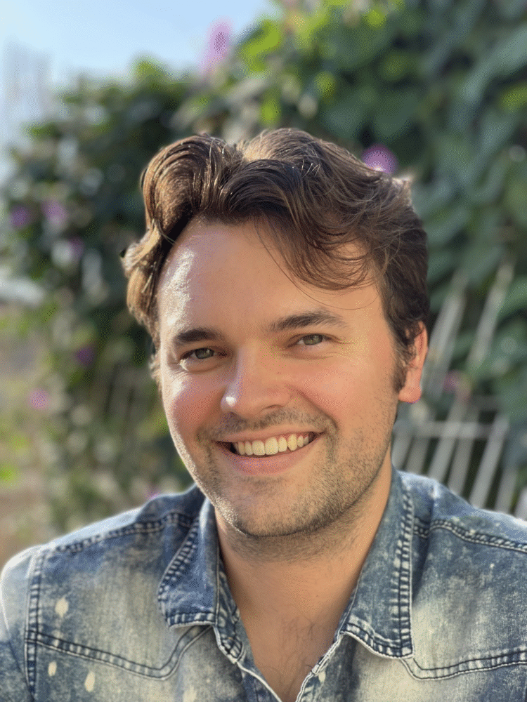 Danny Wagner smiling outdoors in a denim shirt, with green foliage in the background.