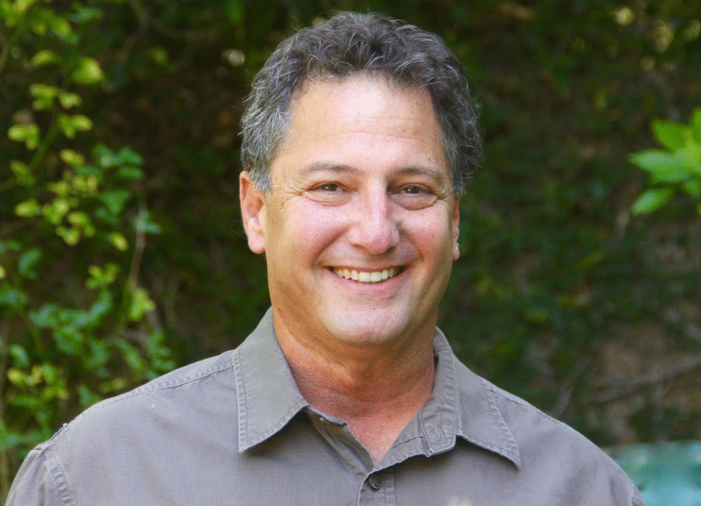 Denny Tedesco, smiling, in a headshot with a leafy green background.