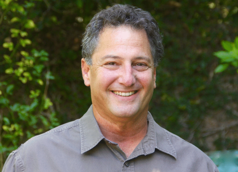 Denny Tedesco, smiling, in a headshot with a leafy green background.