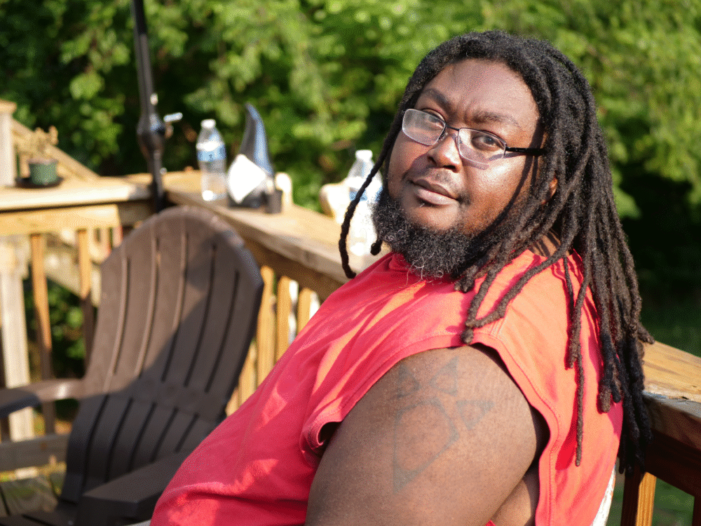 Edward Belcher sitting on a wooden deck with dreadlocks and a red shirt.