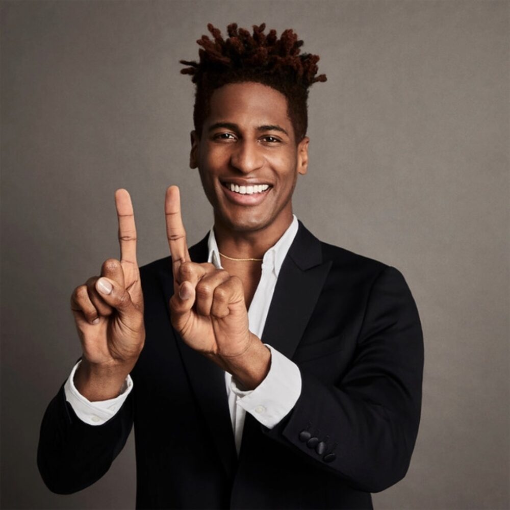 Jon Batiste smiling and holding up two fingers in a black suit.