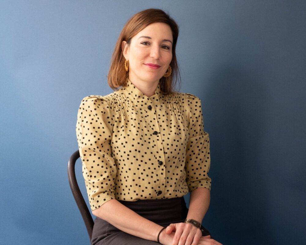 Julia Pontecorvo, smiling, sits against a blue backdrop. She wears a patterned blouse and gold earrings.