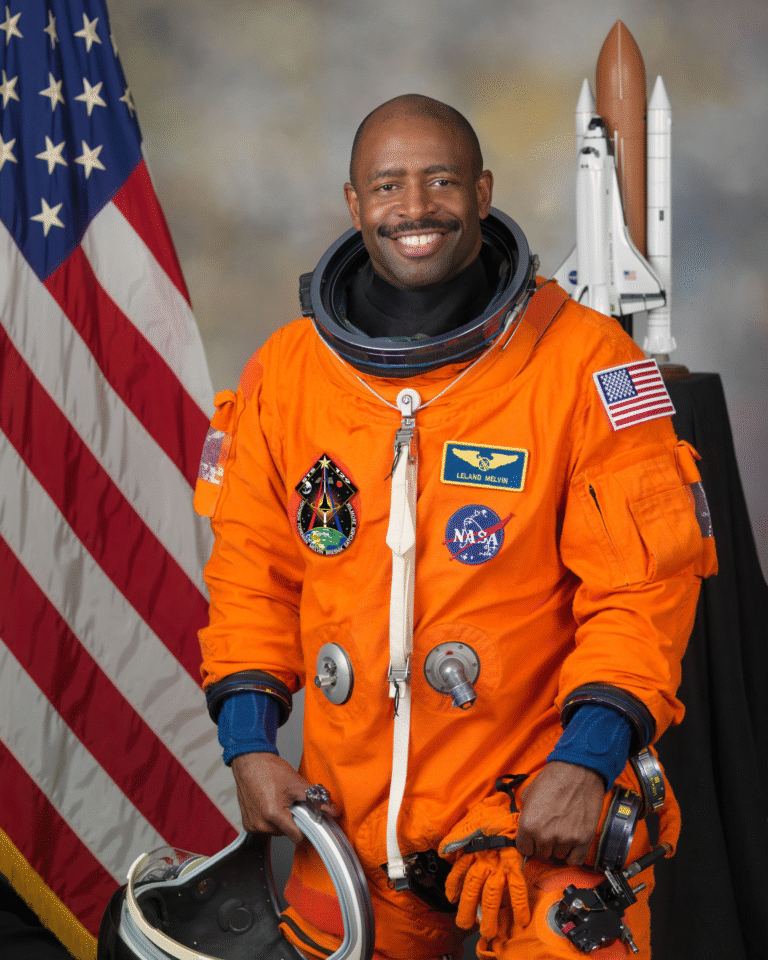 Leland Melvin in an orange spacesuit, smiling with the American flag behind him.