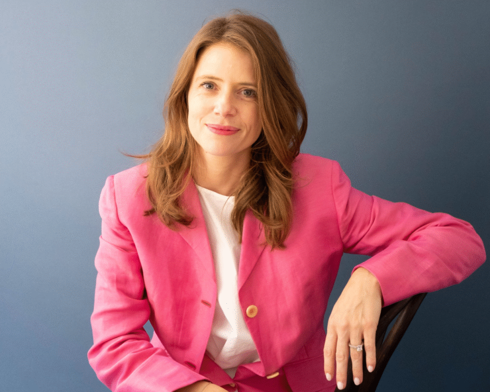 Rachael Dyer, wearing a pink blazer, poses confidently against a blue background.