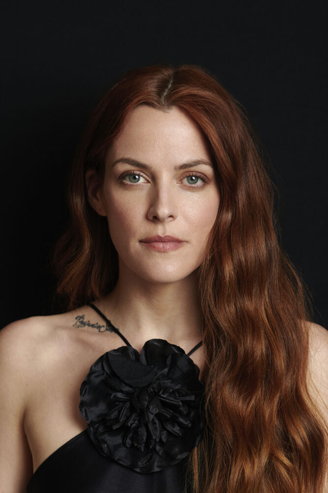 Riley Keough portrait with red hair and a black floral dress against a dark background.