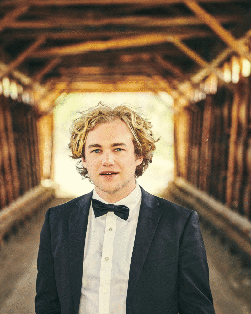 Shaun Couture in a tuxedo stands in a covered bridge with a confident gaze.