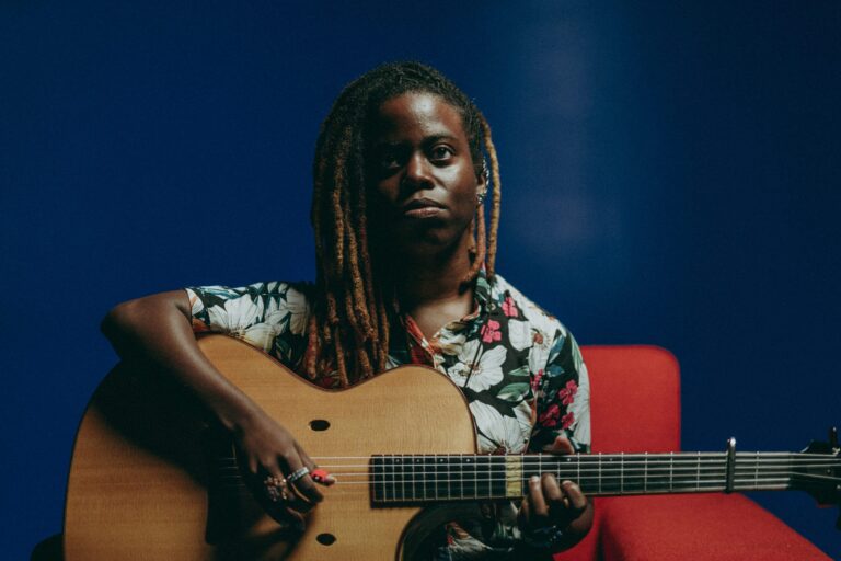 Yasmin Williams playing acoustic guitar, dreadlocks visible, against a blue background.