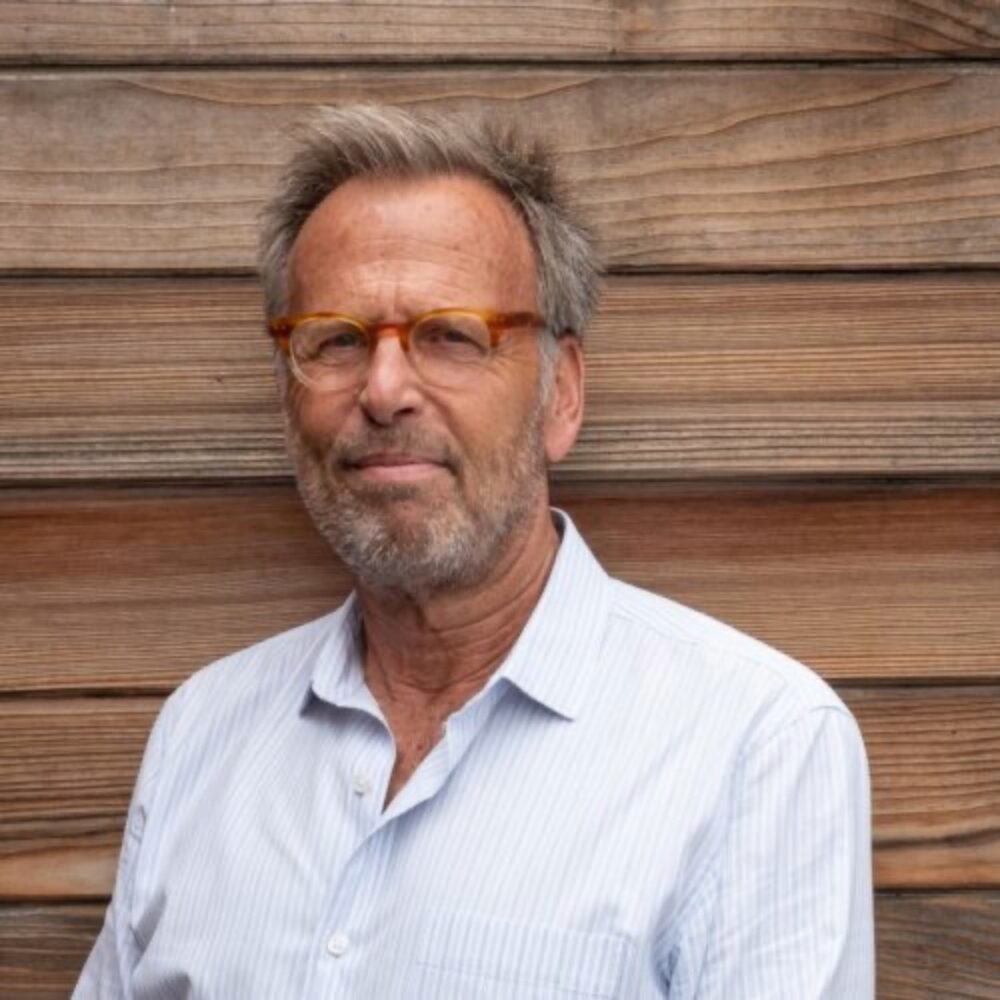 Portrait of Mark Johnson, wearing glasses and a light blue shirt, against a wooden wall.