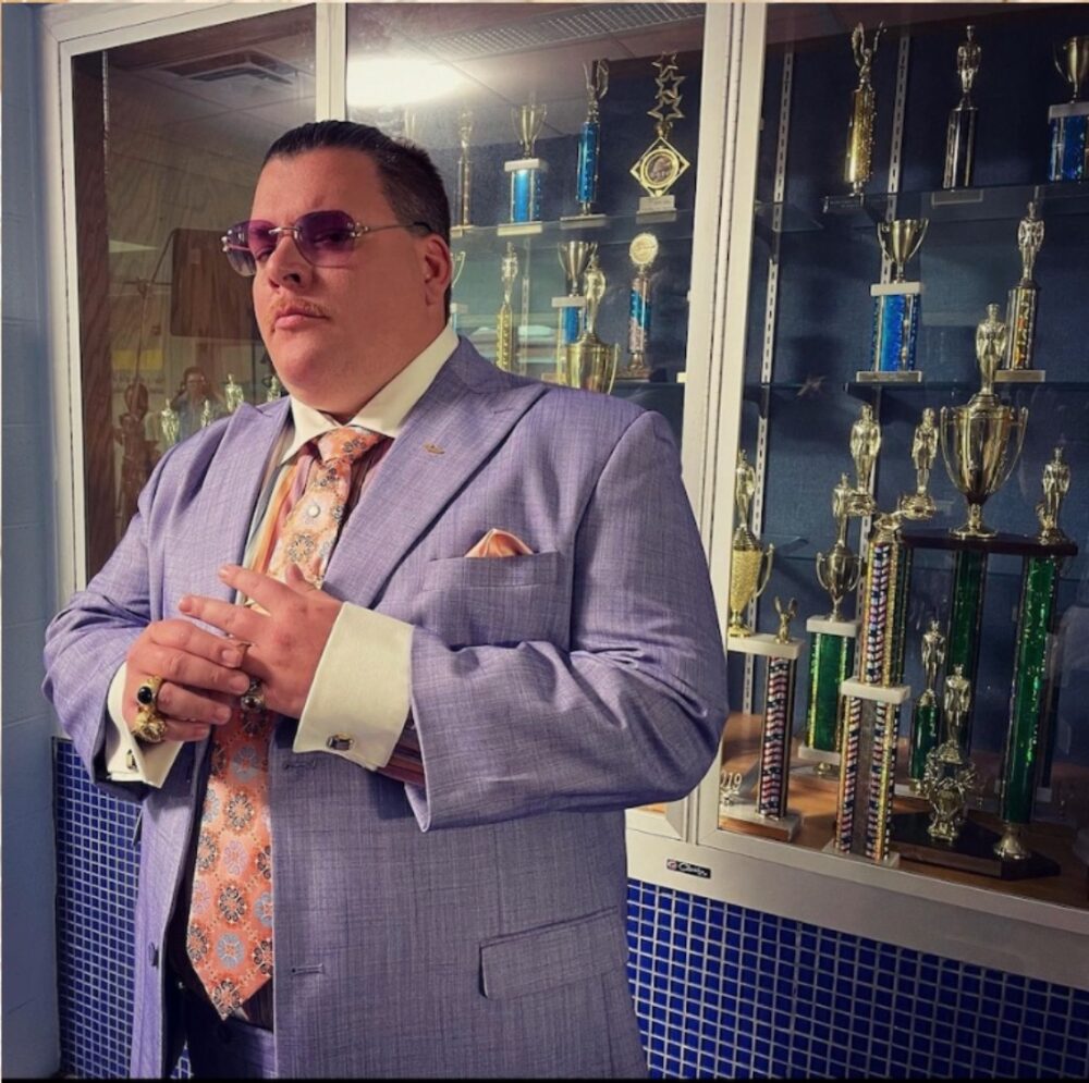 Chad Dukes in a purple suit stands by a trophy case.