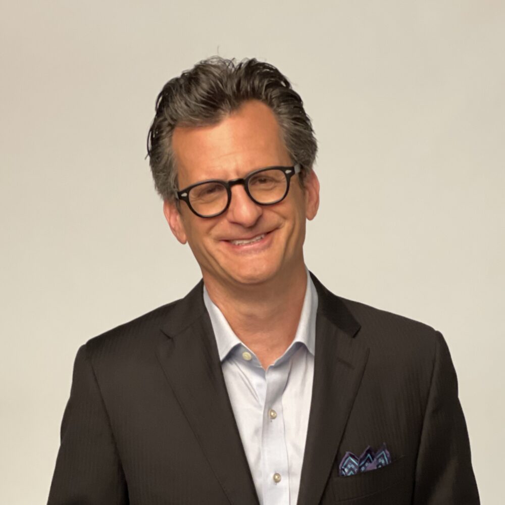 Ben Mankiewicz smiles in a suit and glasses