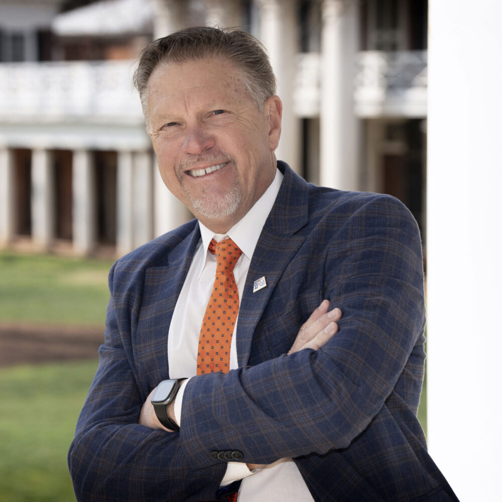 Smiling man in a suit with arms crossed, standing in front of a building.