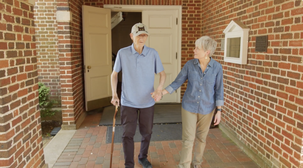 Elderly couple holding hands, walking outside a brick building.