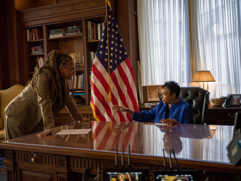 Two women in an office, one standing and one seated at a desk, with an American flag behind them.