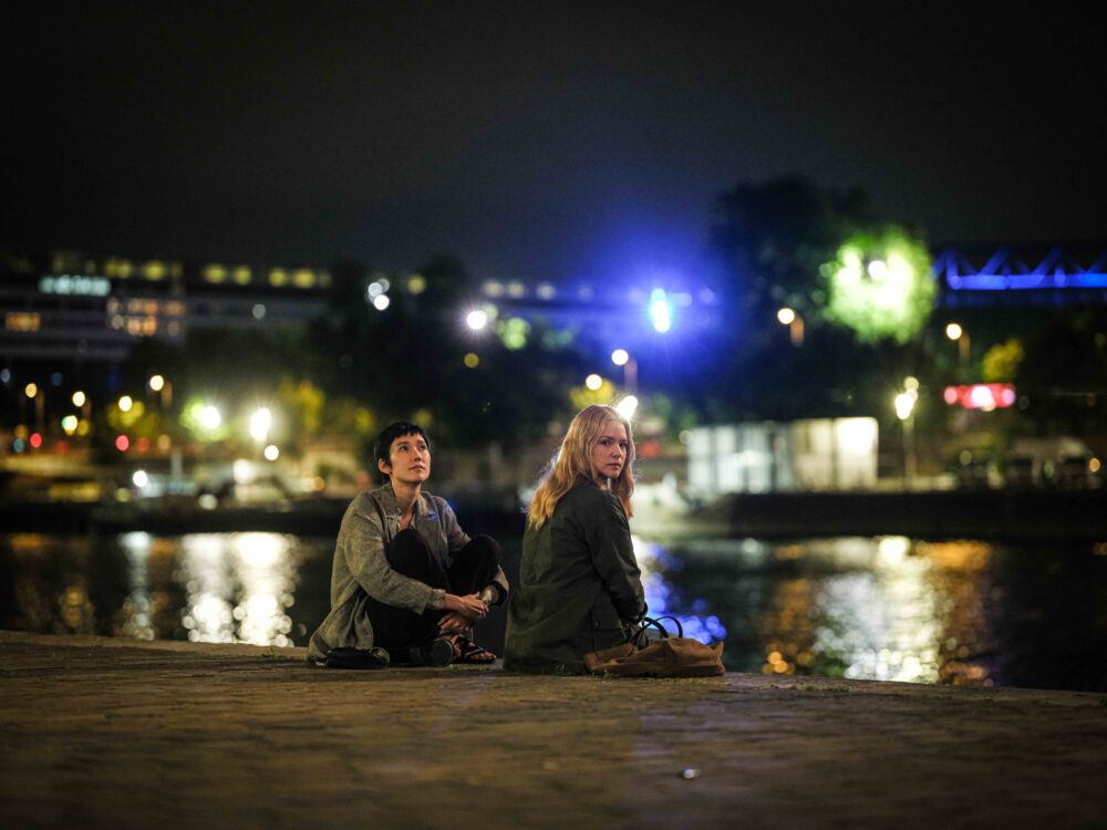 Two women sit by a river at night, looking thoughtful