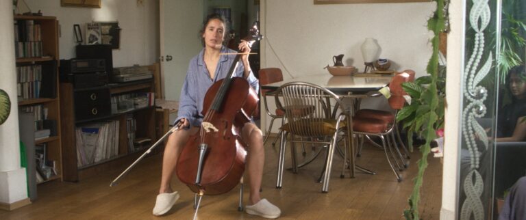Woman with cello in a living room, surrounded by books and plants.