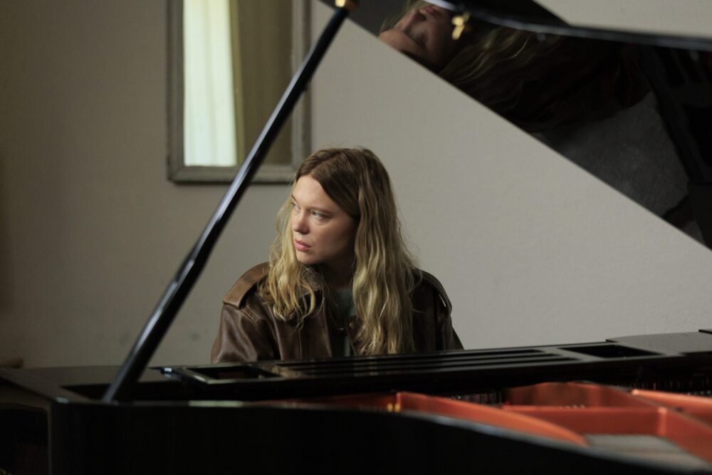 Léa Seydoux plays piano in a dimly lit room, wearing a brown leather jacket.