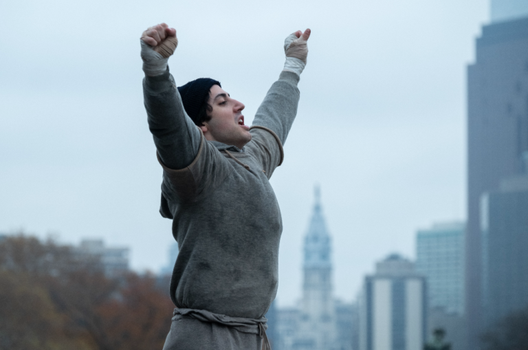 Actor playing Rocky Balboa with arms raised in triumph.
