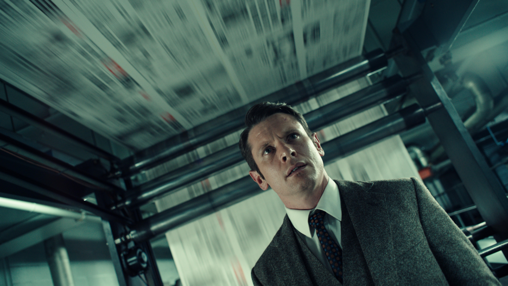 Man in suit looks up at printing press with blurred newspaper sheets