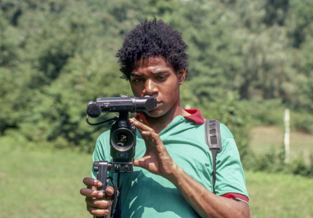 Young Jean-Michel Basquiat holds a video camera outdoors.
