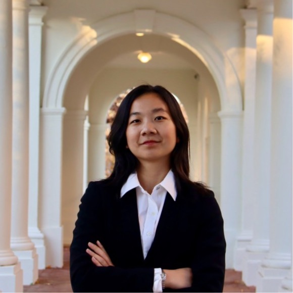 Olivia Zheng, a young Asian woman in a black suit and white shirt, stands with arms crossed.