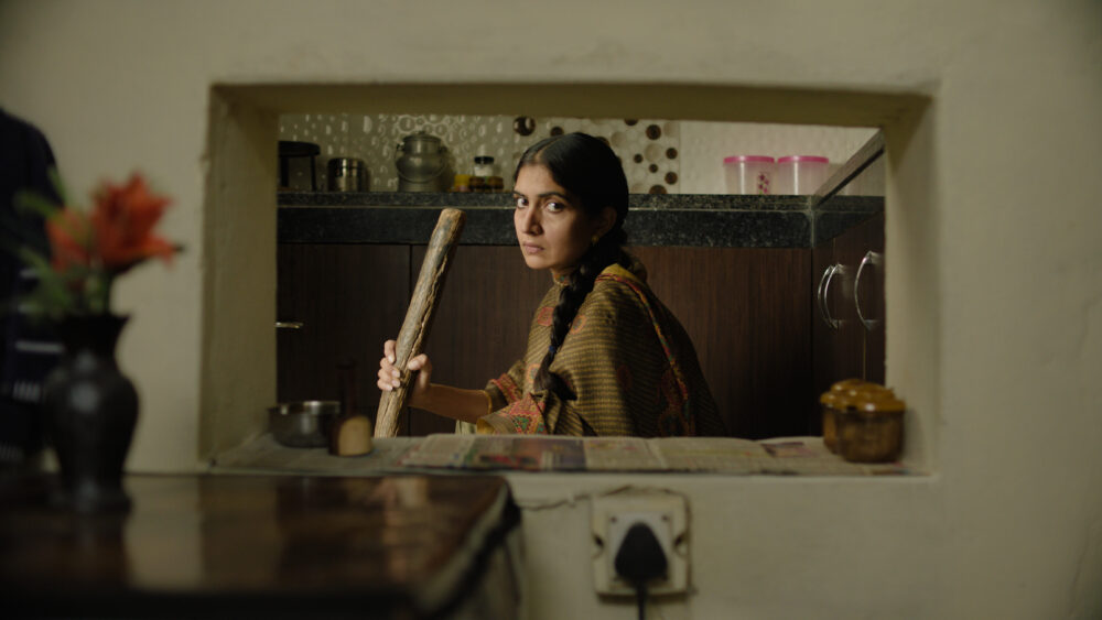Rehmat, a young woman in traditional attire, holds a wooden stick in a kitchen.