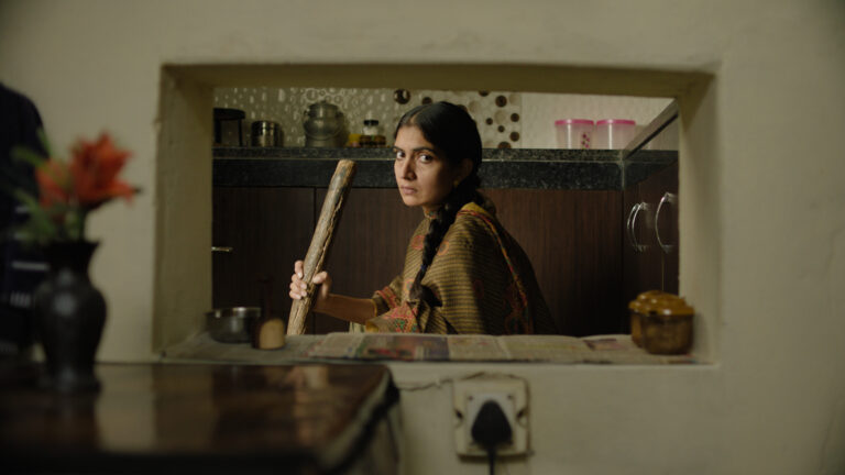 Rehmat, a young woman in traditional attire, holds a wooden stick in a kitchen.