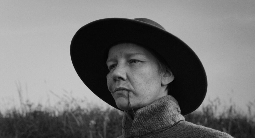 Close-up of a woman in a wide-brimmed hat, with a piece of straw hanging from her mouth.
