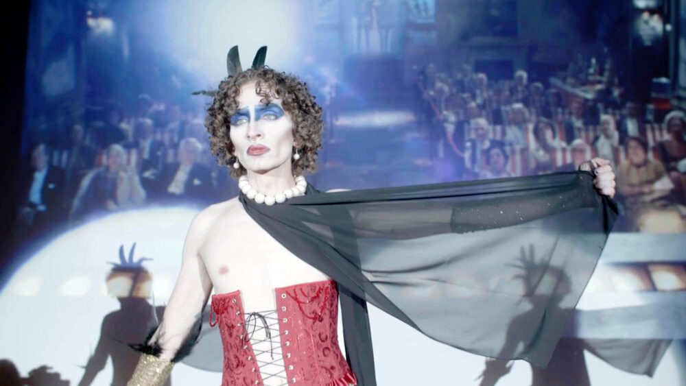 Performer in red corset and blue makeup on stage, "Time Warp" theme.