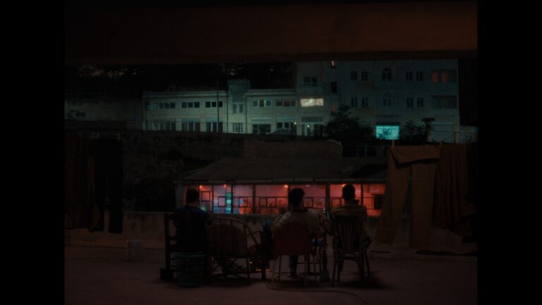 Three figures sit on a rooftop at night, facing a building with colorful lights.