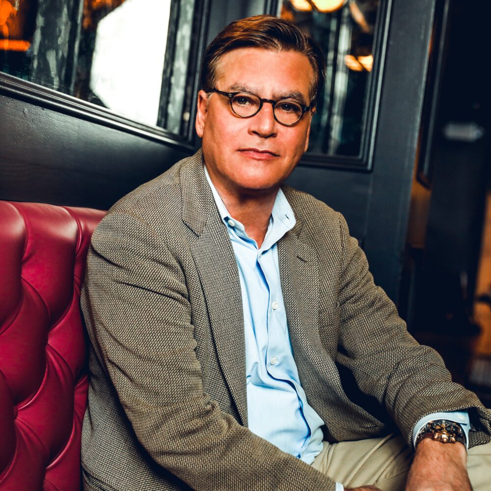 Aaron Sorkin in a tweed jacket and glasses, seated indoors.