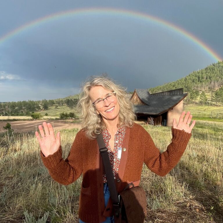 Betsy Kenyon smiles with arms raised under a rainbow in a grassy field.
