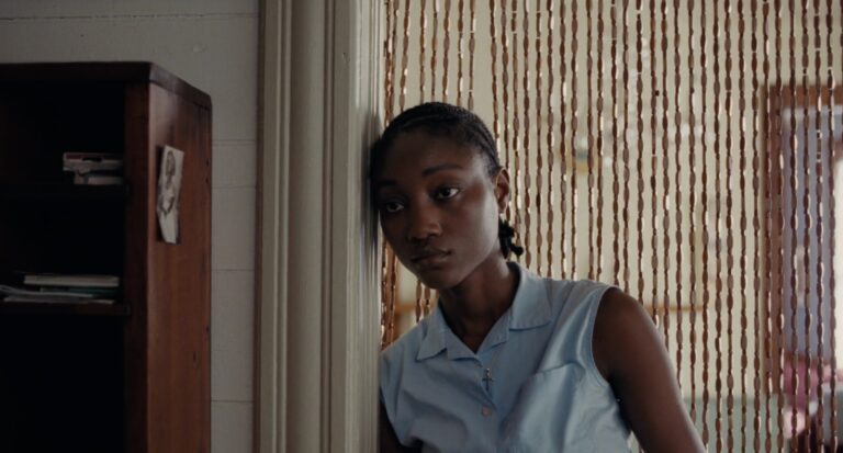 Young Black woman in a blue shirt leans against a doorframe, looking thoughtfully.