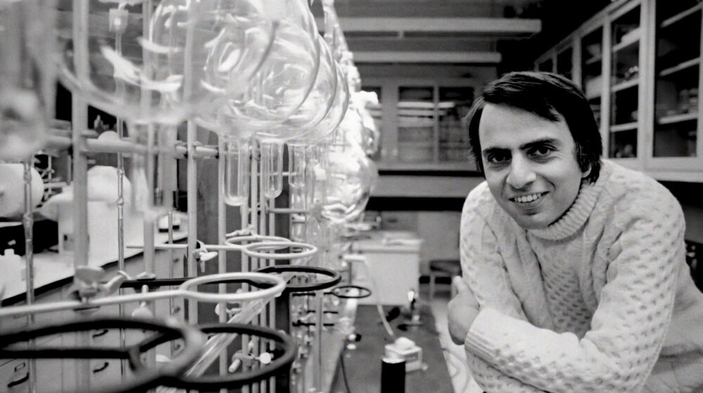 Carl Sagan smiles in a lab with glassware, embodying "We Are Made of Star Stuff.