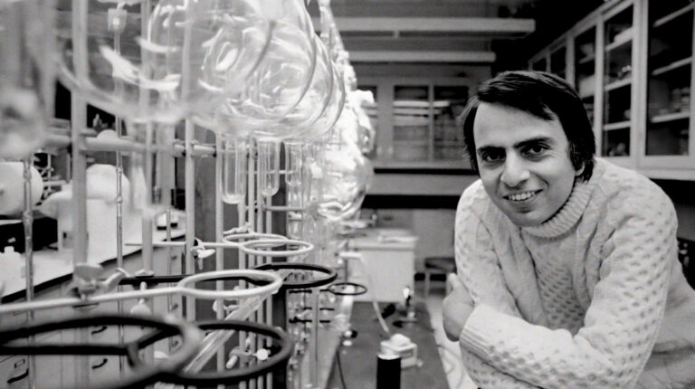 Carl Sagan smiles in a lab with glassware, embodying "We Are Made of Star Stuff.