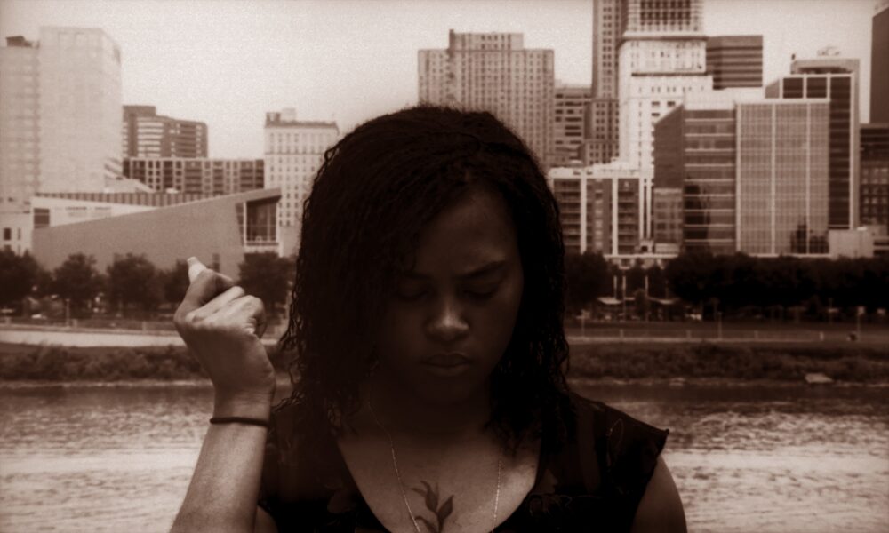 Woman with braided hair looks down in front of a city skyline, a Kevin Jerome Everson film still.