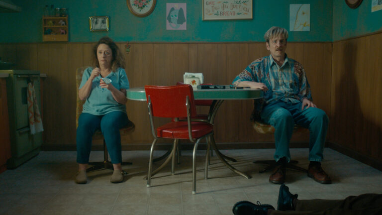 Couple sitting at a retro diner table, one eating, one looking at camera
