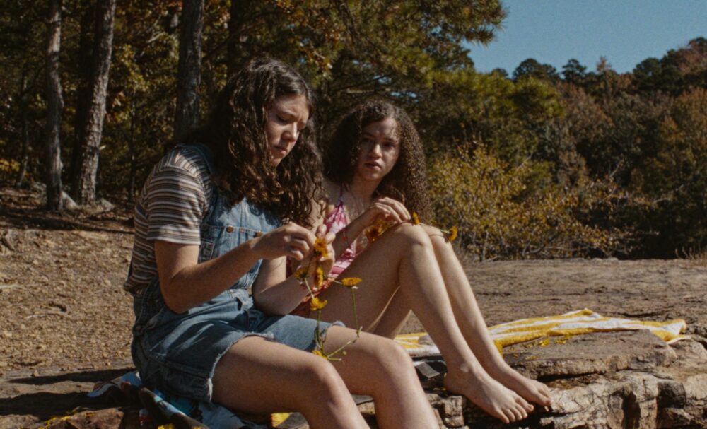 Two young girls sit on rocks, one picking flowers, in a natural outdoor setting.