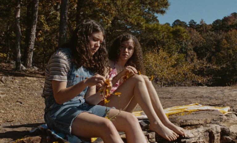 Two young girls sit on rocks, one picking flowers, in a natural outdoor setting.