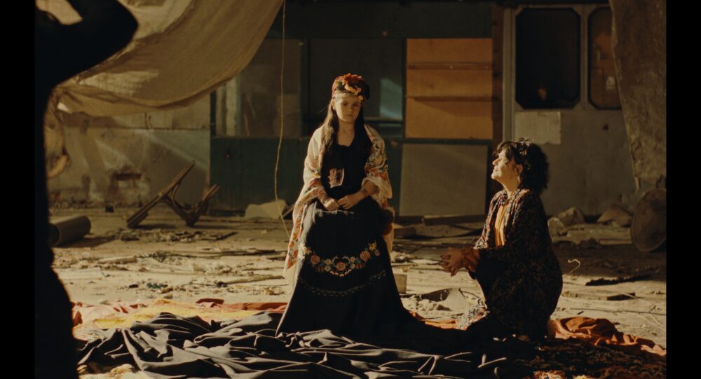Nina Roza: Woman in ornate dress and flower crown kneels before seated woman.