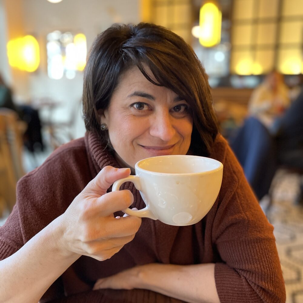 Pam Hervey smiles, holding a white coffee mug in a cafe.