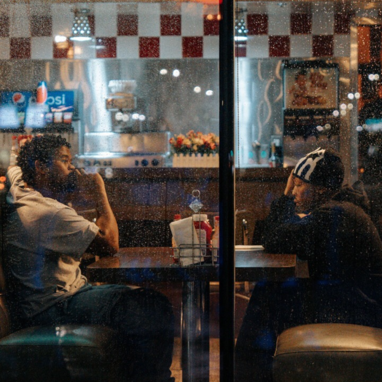 Two men sit at a diner table on a rainy night, reflecting on 