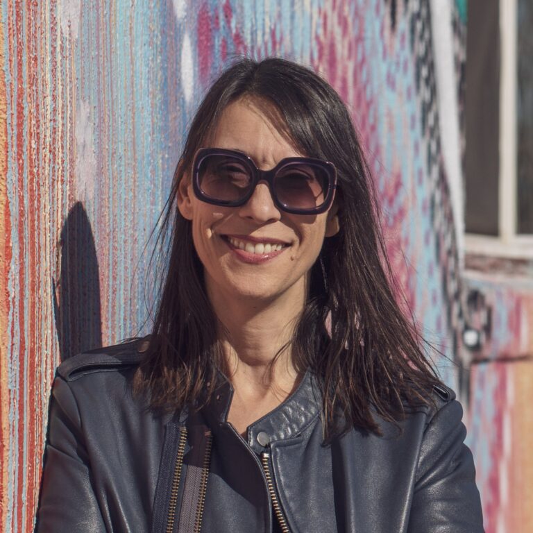 Sheryl Glubok smiles in front of a colorful graffiti wall.
