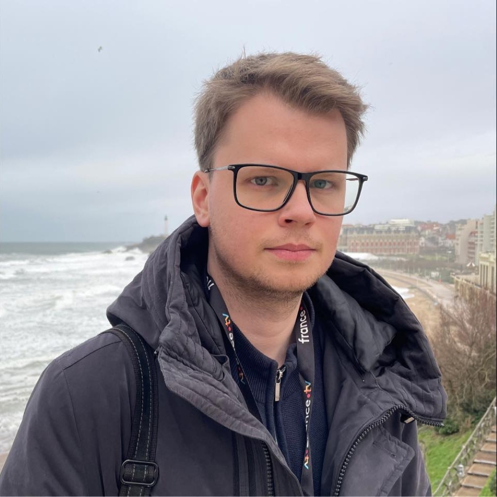 Tomáš Gič in a dark jacket with a FranceTV lanyard, overlooking a coastline.