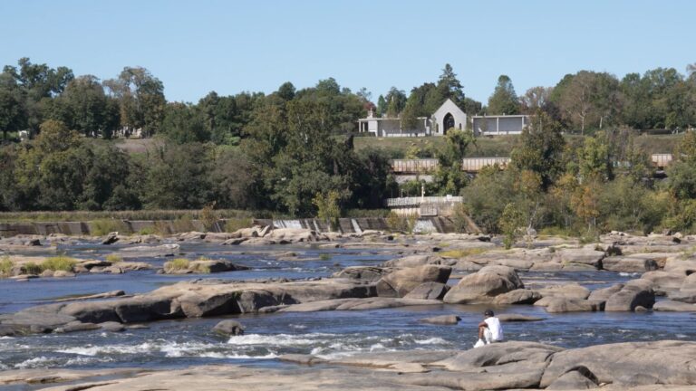 Man sits on rocks in a river with a dam and buildings in the background