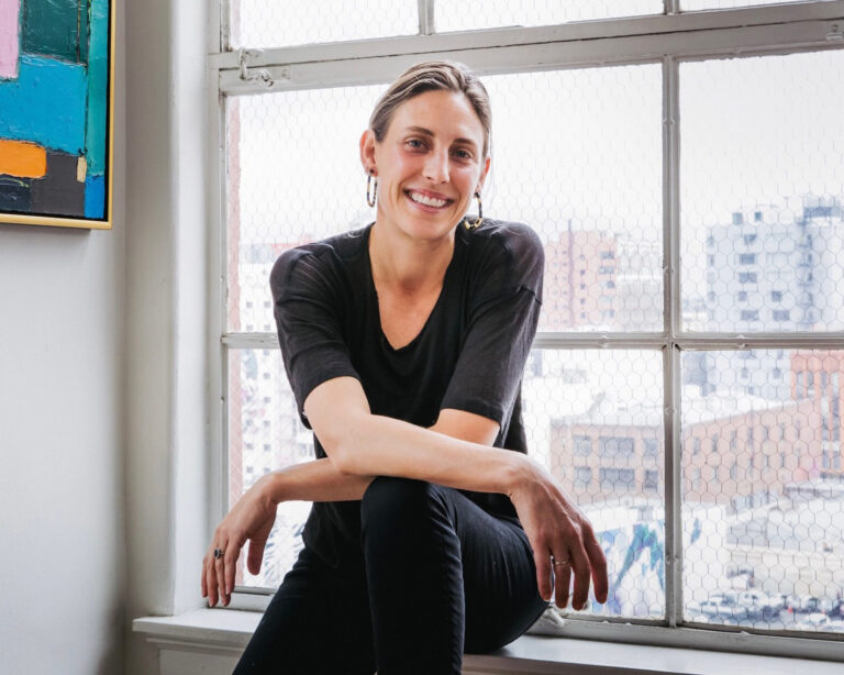 Smiling woman in black sitting by a large window with city buildings outside