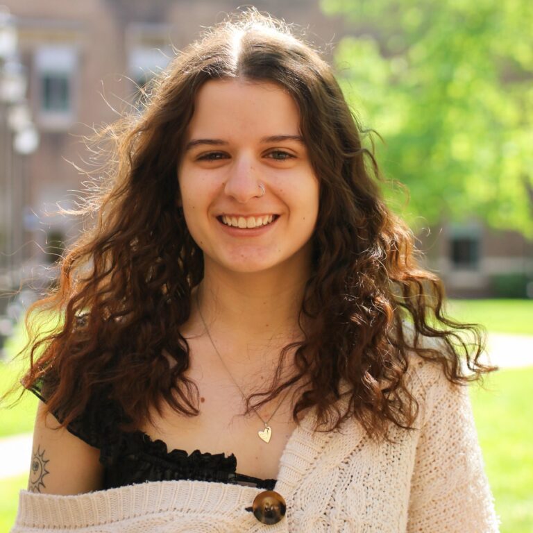Caroline Crain smiling outdoors with curly brown hair and a heart necklace.