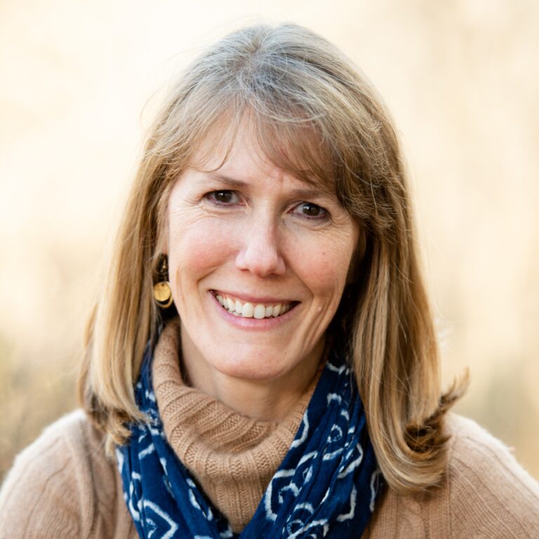 Elizabeth Shillue smiles warmly, wearing a tan sweater and blue scarf.